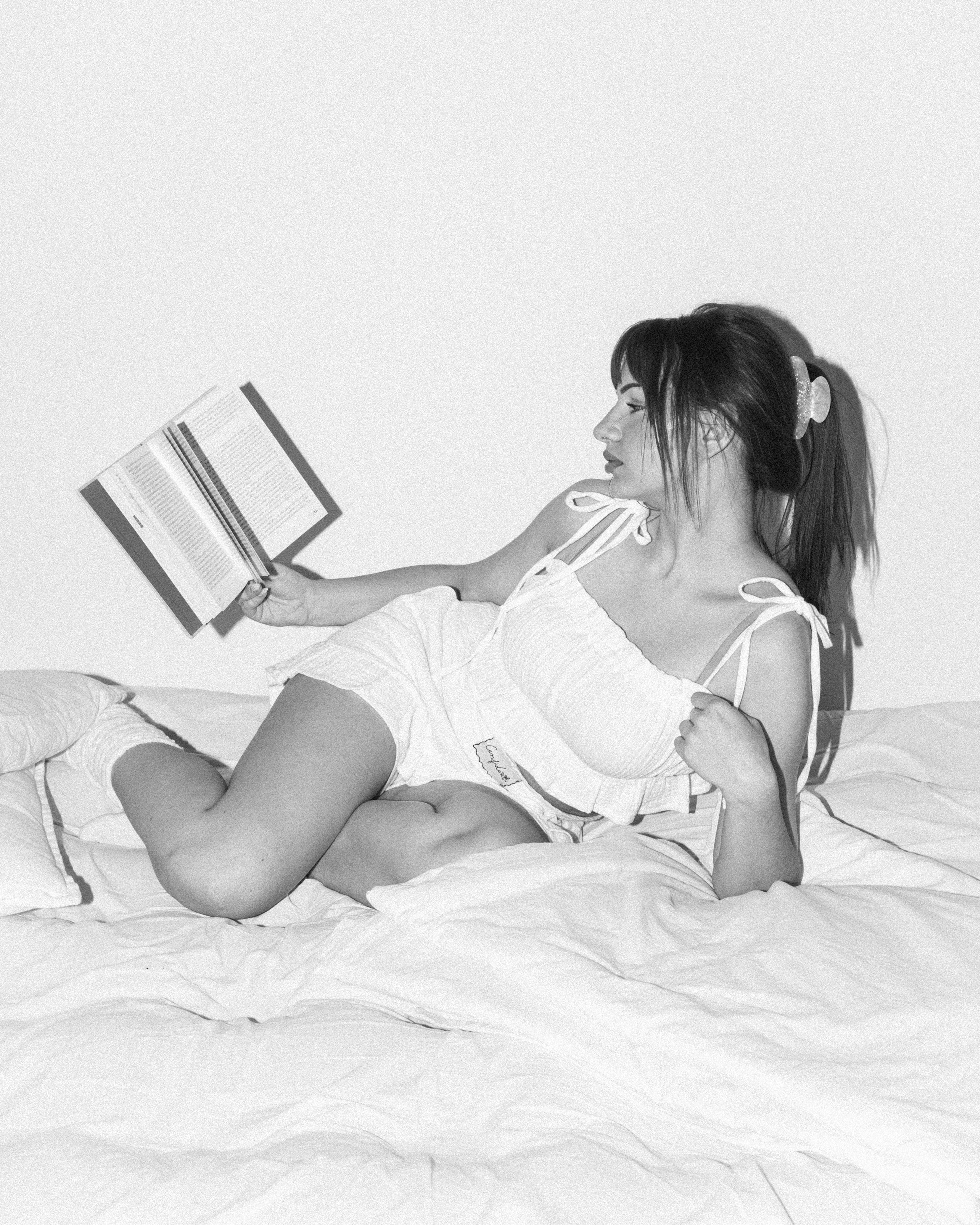 ALT: Dámský mušelínový set Luna Comfident Studio, bílé šortky s elastickým pasem a top na ramínka se zavazováním, lifestyle foto ženy ležící na posteli s knihou, černobílá fotografie