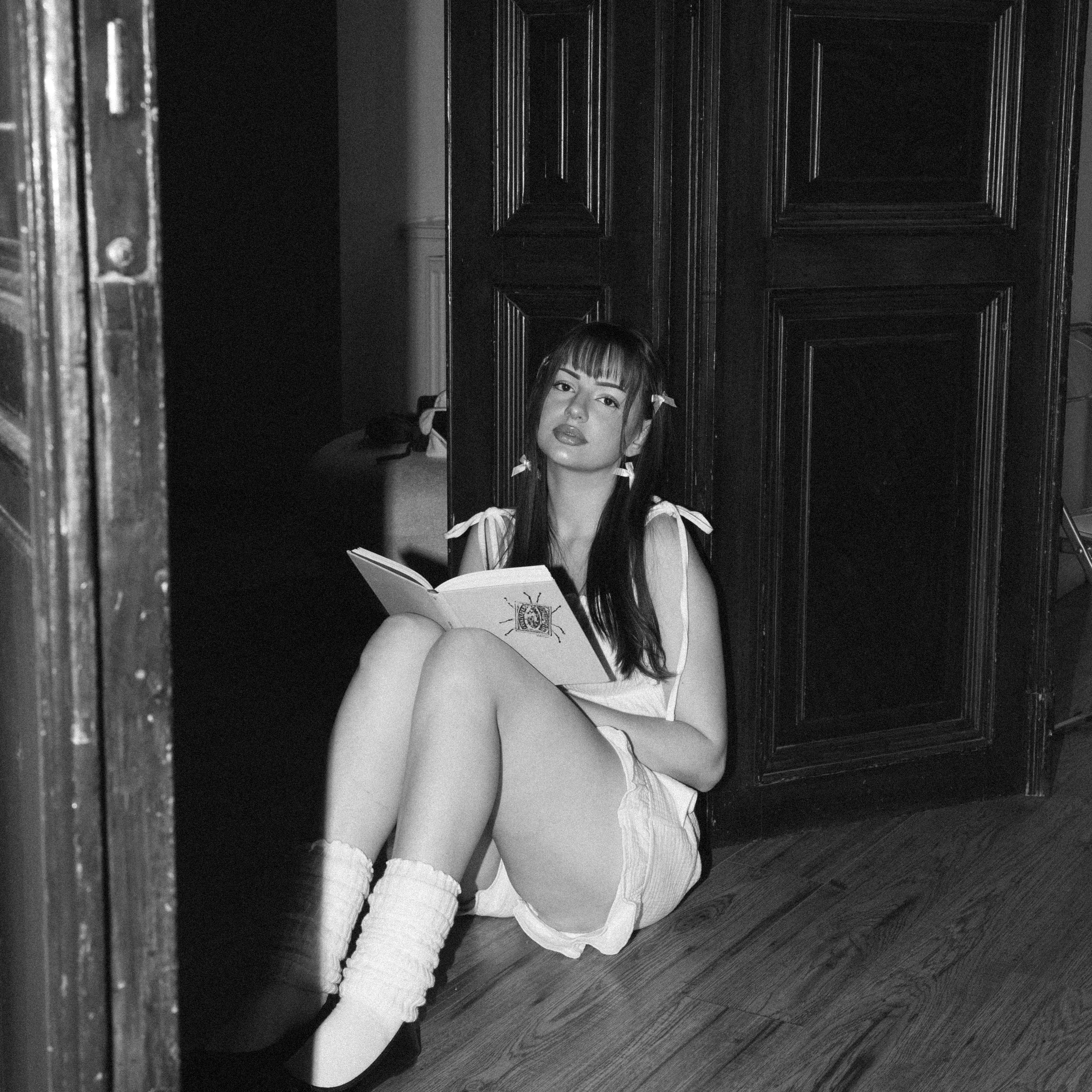 Dámský mušelínový set Luna Comfident Studio, bílé šortky a top na ramínka, lifestyle foto ženy sedící na podlaze u dřevěných dveří s knihou, černobílá fotografie
